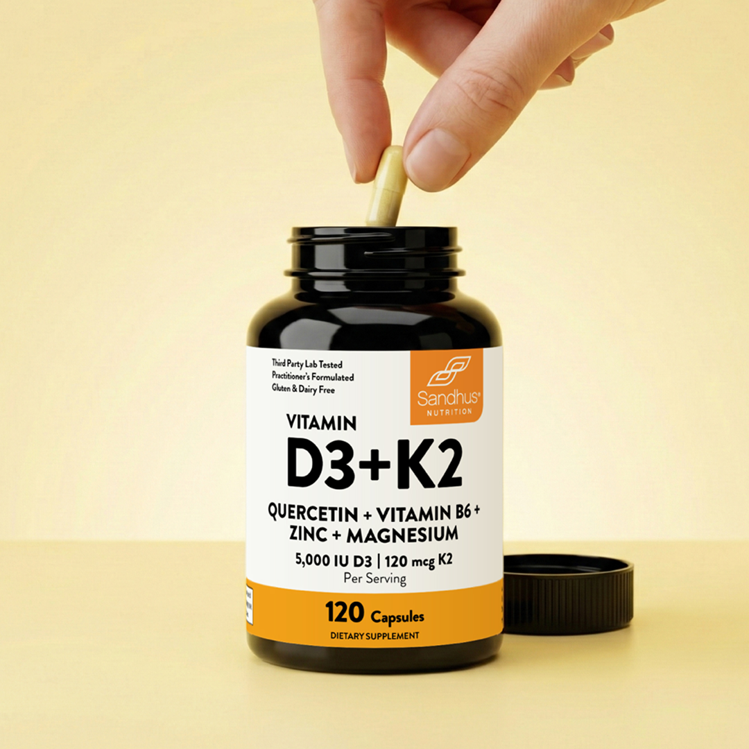 A hand places a capsule into an open bottle of Sandhus Nutrition Vitamin D3+K2 supplements.