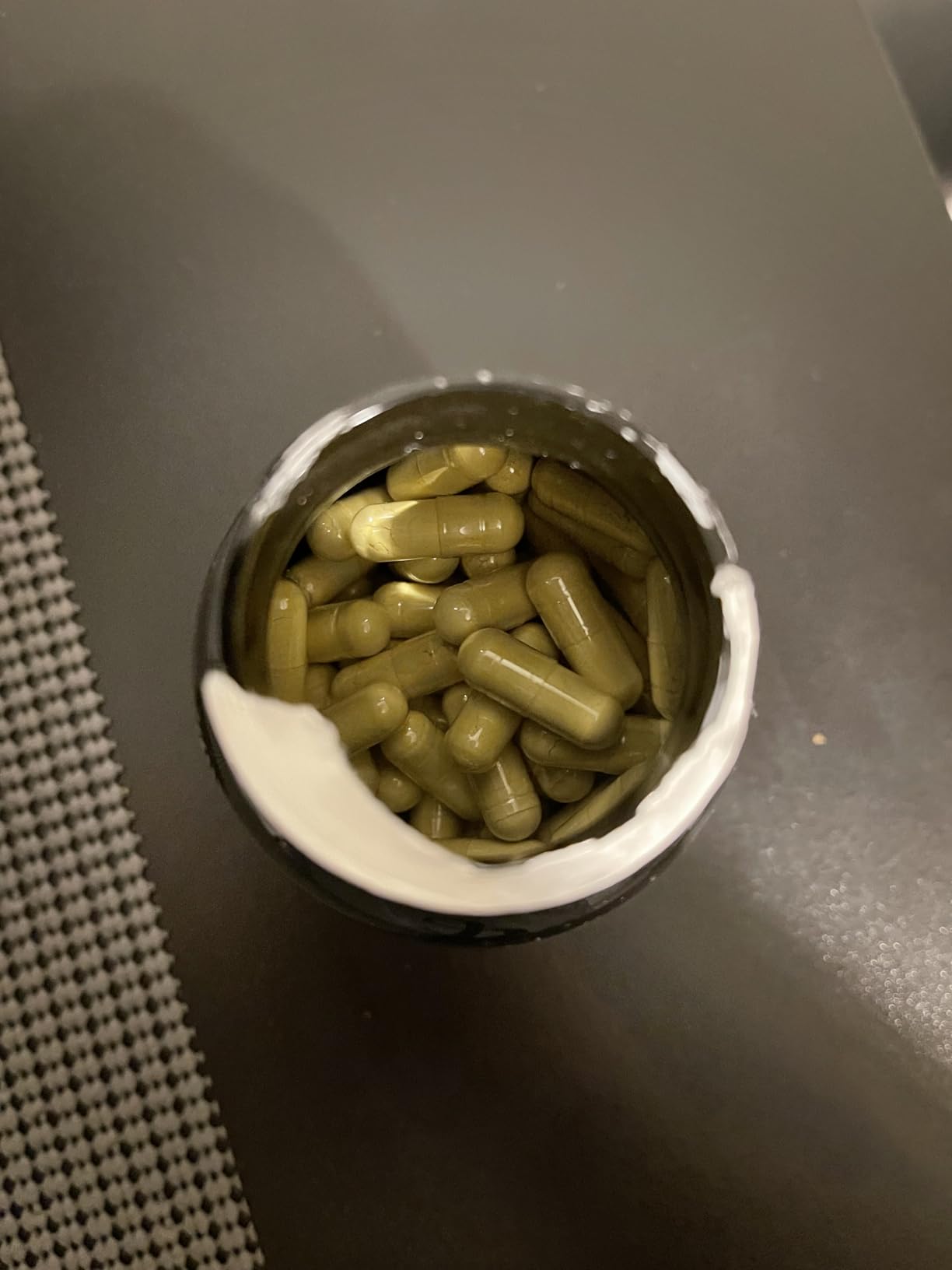 Open bottle filled with green capsules on a dark surface.
