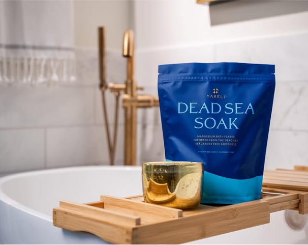 Blue Dead Sea soak package and gold candle on a wooden bath tray.