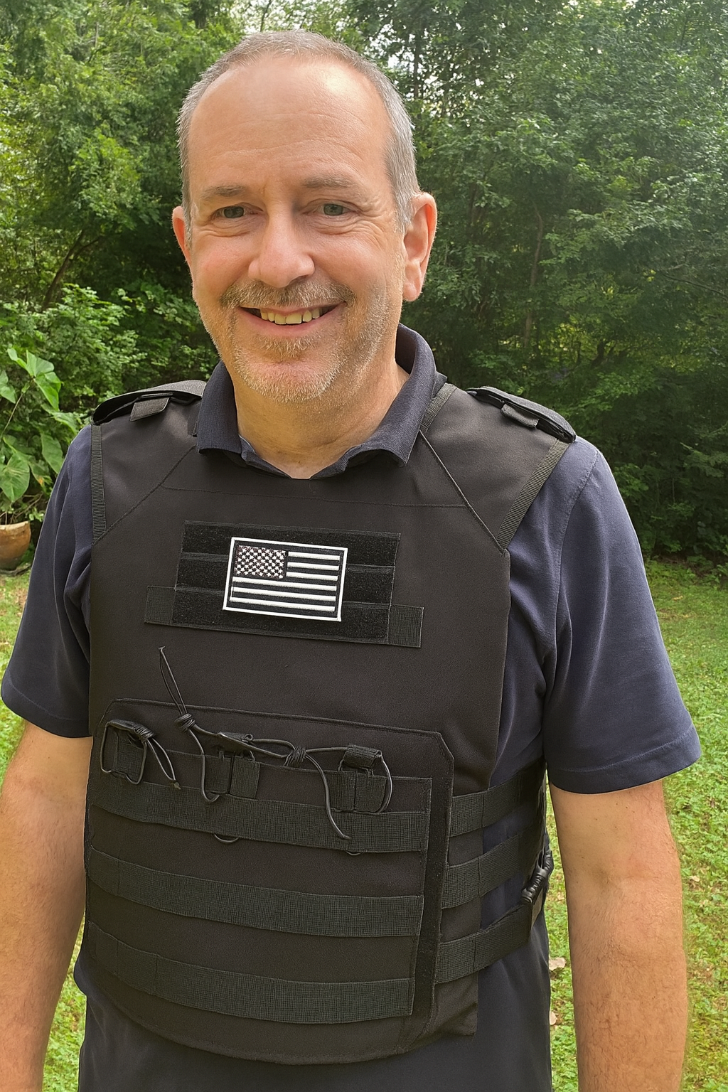 Smiling person in a black tactical vest with an American flag emblem, outdoors.