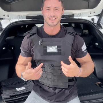 Man wearing tactical vest with flag emblem sitting in car trunk.