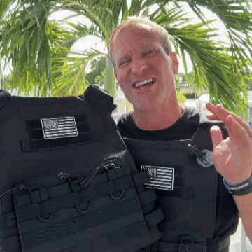 Man holding body armor vest with American flag patch, standing in front of palm trees.