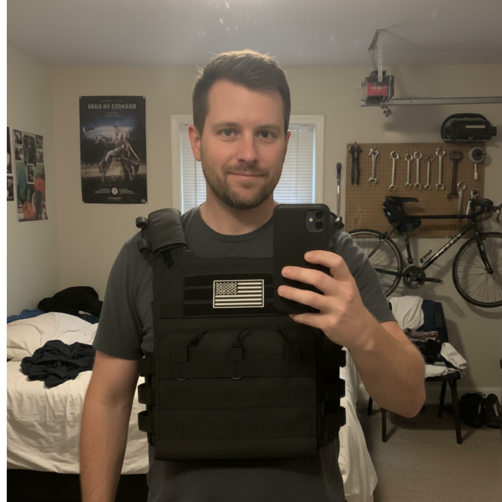 Person taking a mirror selfie wearing a tactical vest in a room with a bicycle and tools on the wall.