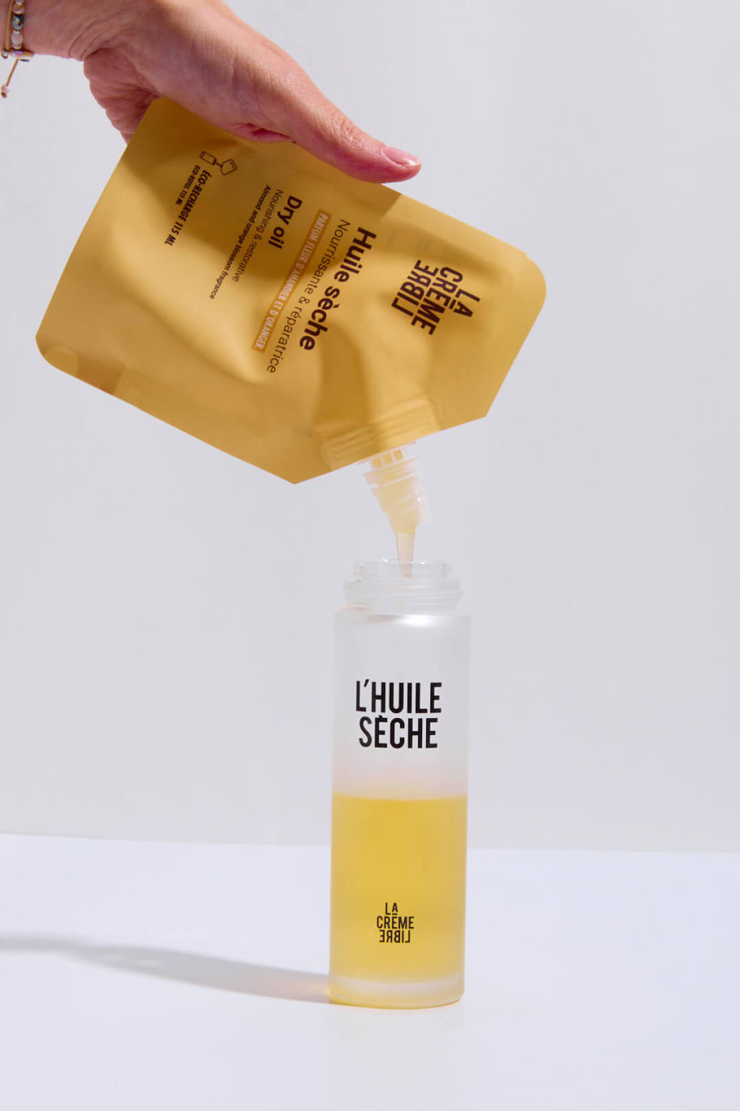 Hand pouring oil from a pouch into a bottle labeled 'L'Huile Sèche.'