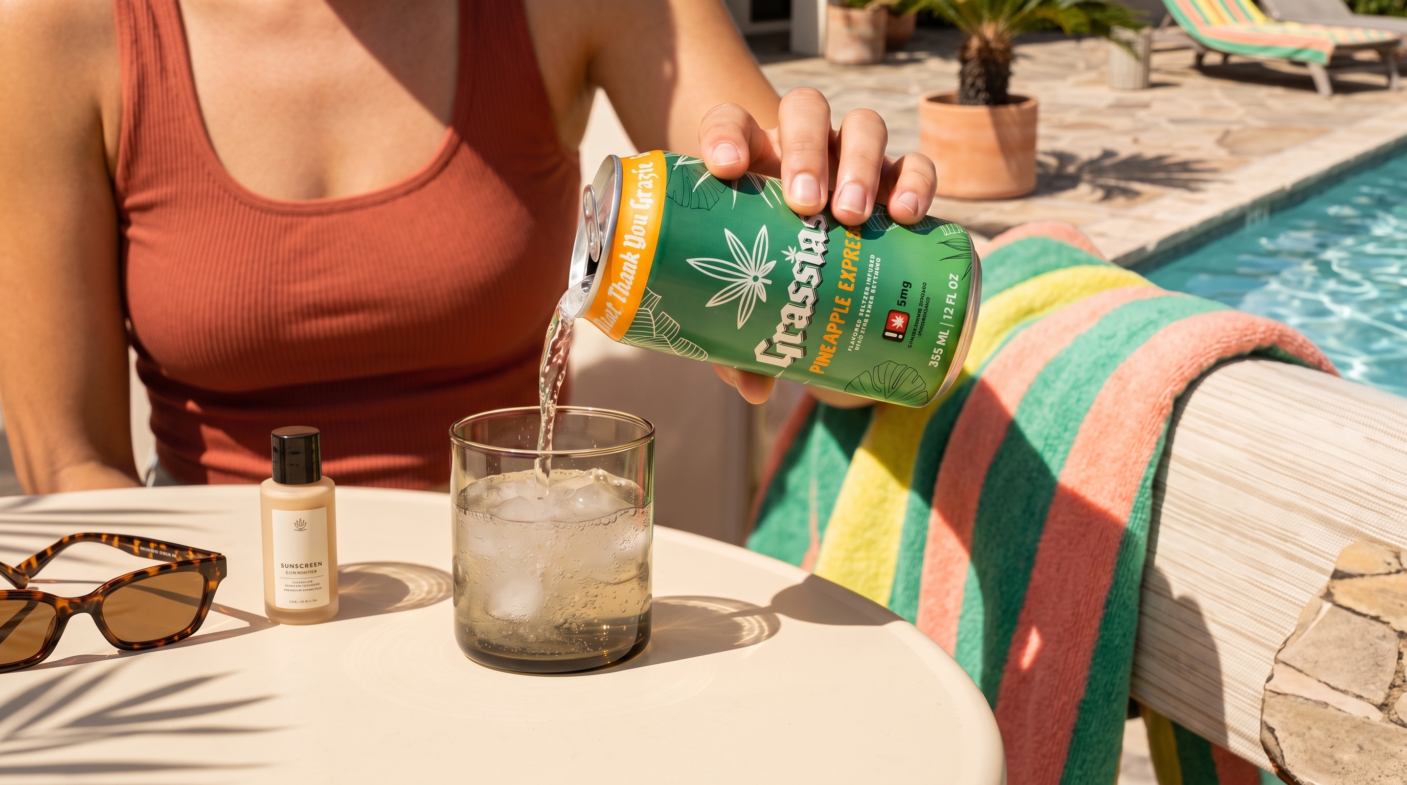 A person pouring a can of Grasstas Pineapple Express seltzer into a glass with ice by a pool.