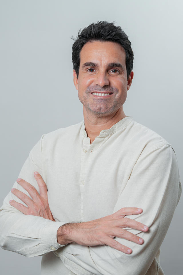 A smiling, dark-haired man in a light-colored shirt stands with his arms crossed against a grey background.