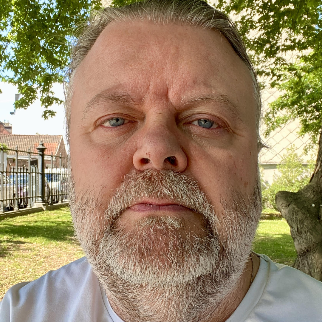 Man with a beard outdoors, trees and buildings in background.