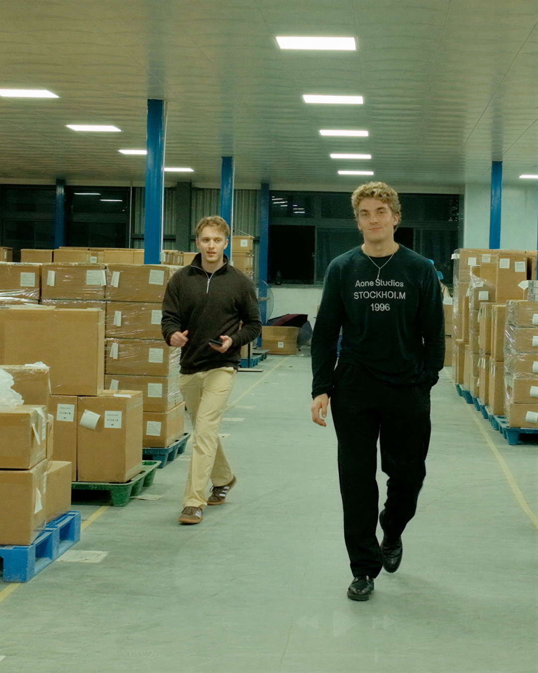 Two people walking in a warehouse with cardboard boxes on pallets.