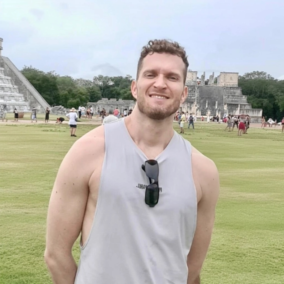 Person smiling in front of ancient ruins and grassy field.
