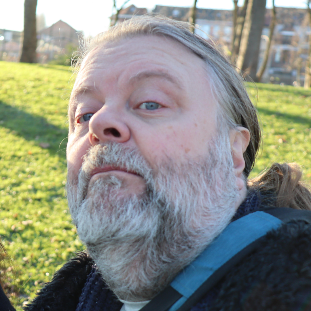 Close-up of a bearded man outdoors with trees and grass in the background.