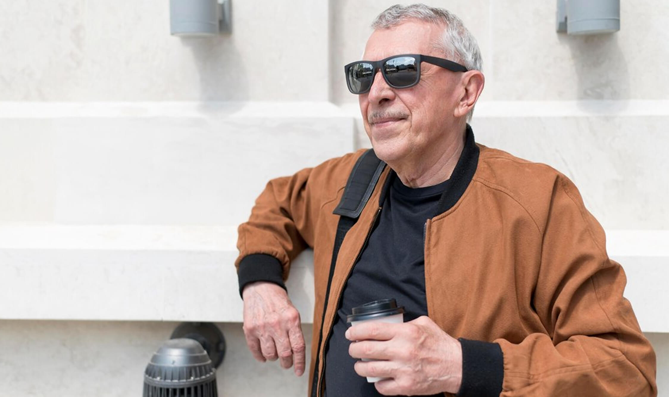 Man in sunglasses and brown jacket holding a coffee cup, leaning against a wall.
