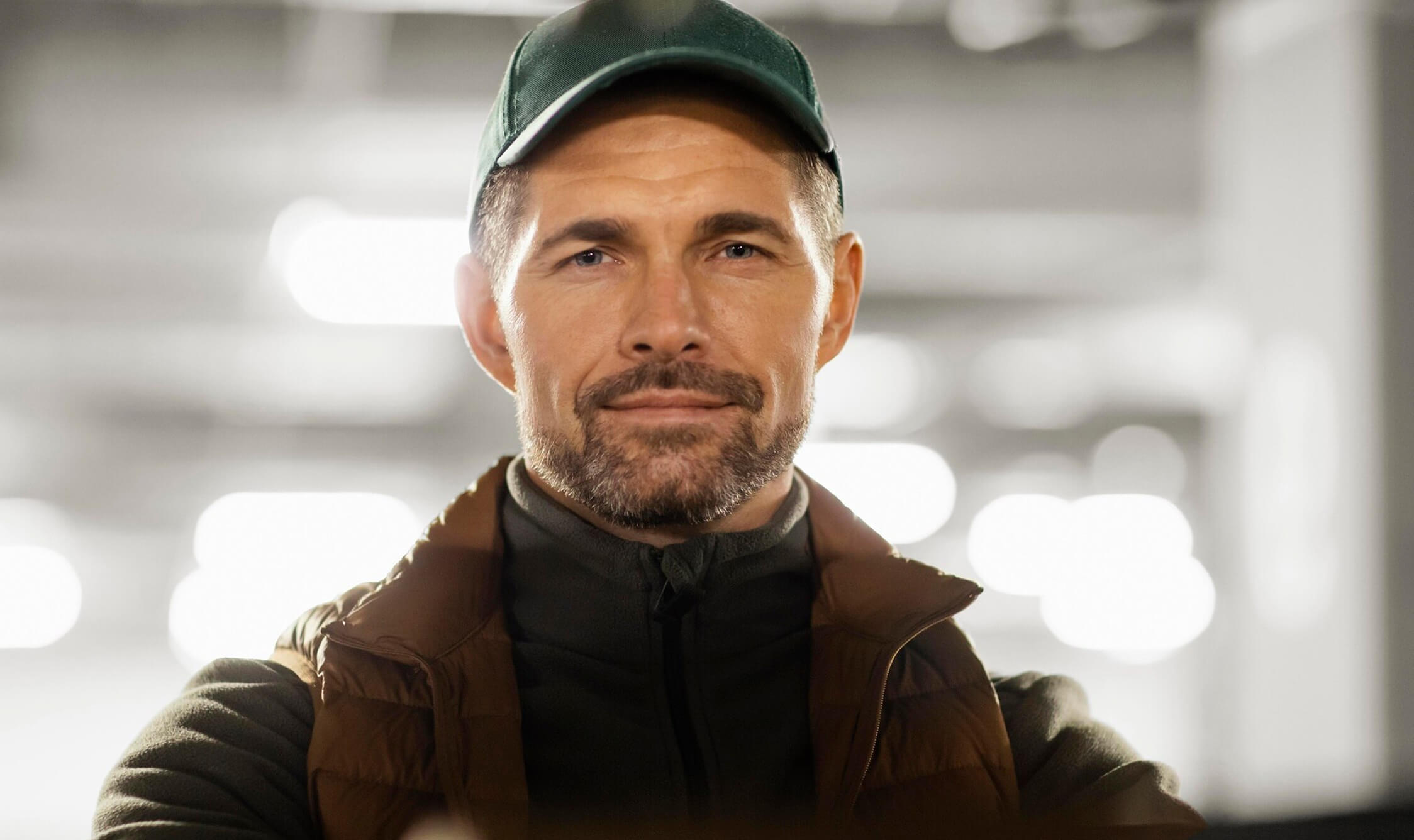 Man wearing a green cap and brown jacket, smiling slightly.