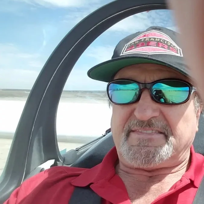 Man wearing sunglasses and a cap, sitting in a vehicle with a beach background.