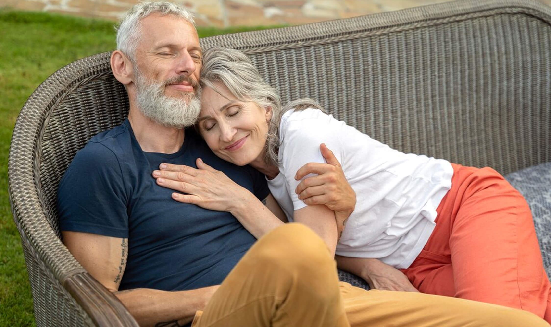 A couple relaxing on a wicker sofa outdoors, smiling with eyes closed.