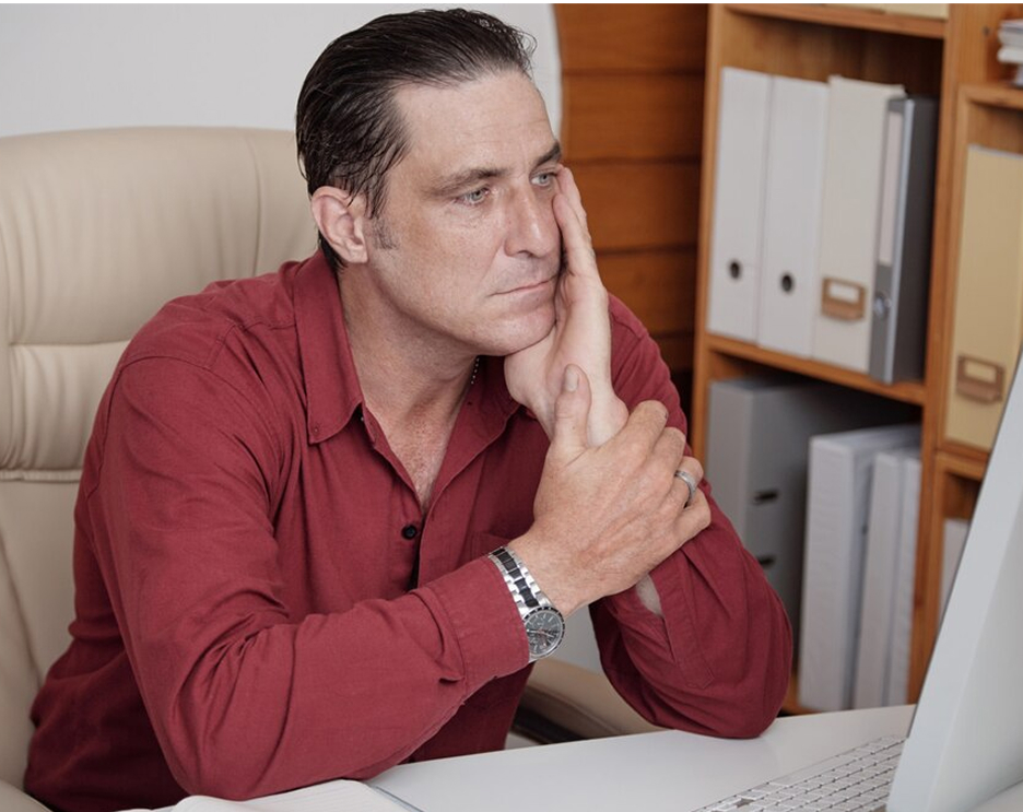 Man in red shirt sitting at a desk, looking at a computer screen.