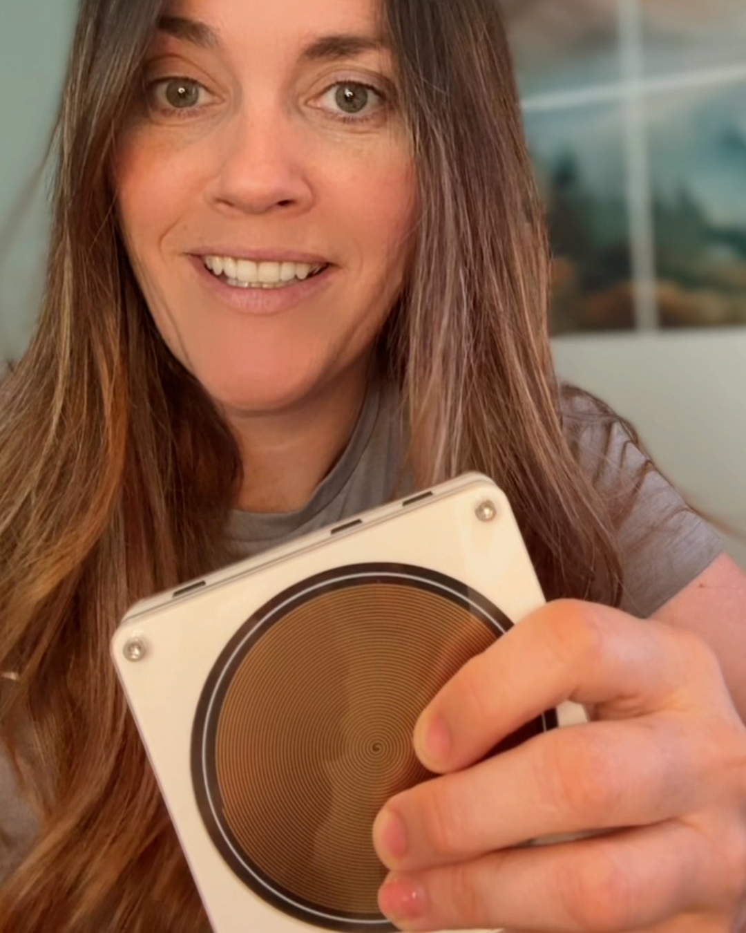A smiling woman with long brown hair holding a square device with a bronze spiral design.