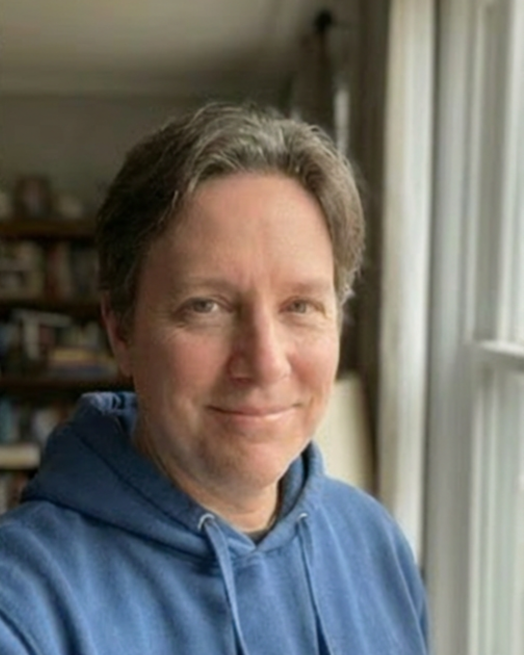 Person in a blue hoodie smiling indoors by a window.