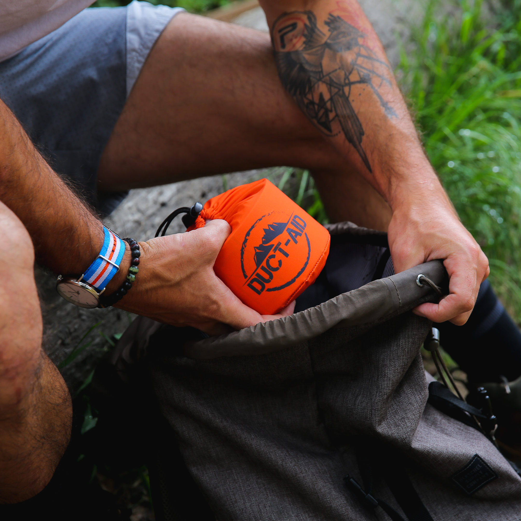 Person placing an orange bag labeled 'DUCT-AID' into a backpack outdoors.