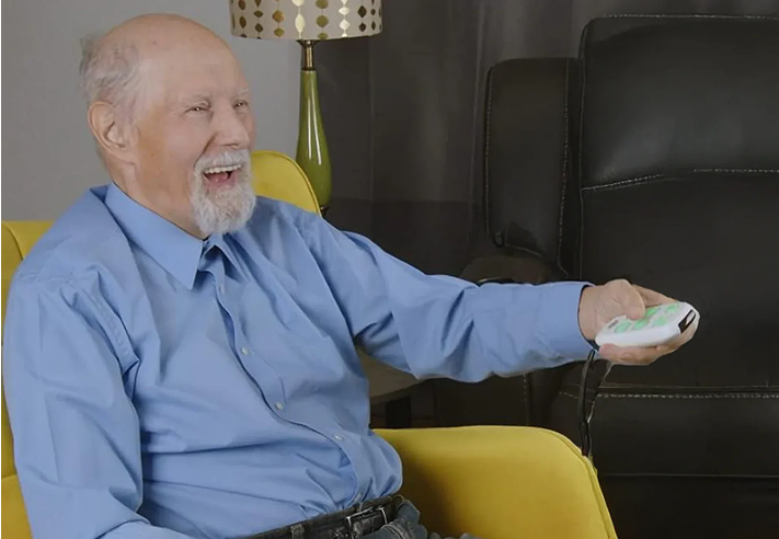Elderly man in blue shirt smiling while holding a remote control.