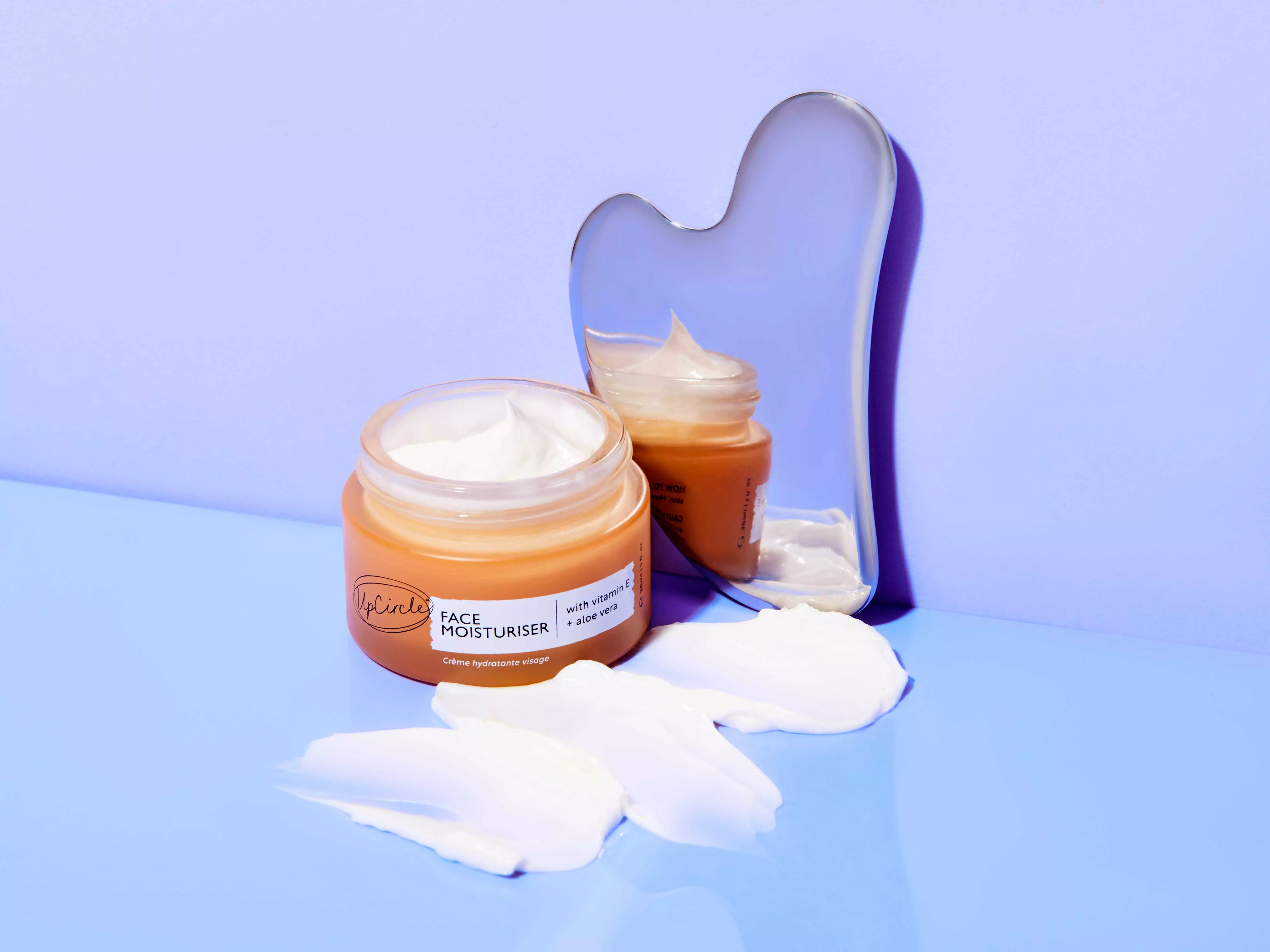 A jar of UpCircle face moisturizer with smears of cream and a reflective gua sha tool on a surface.