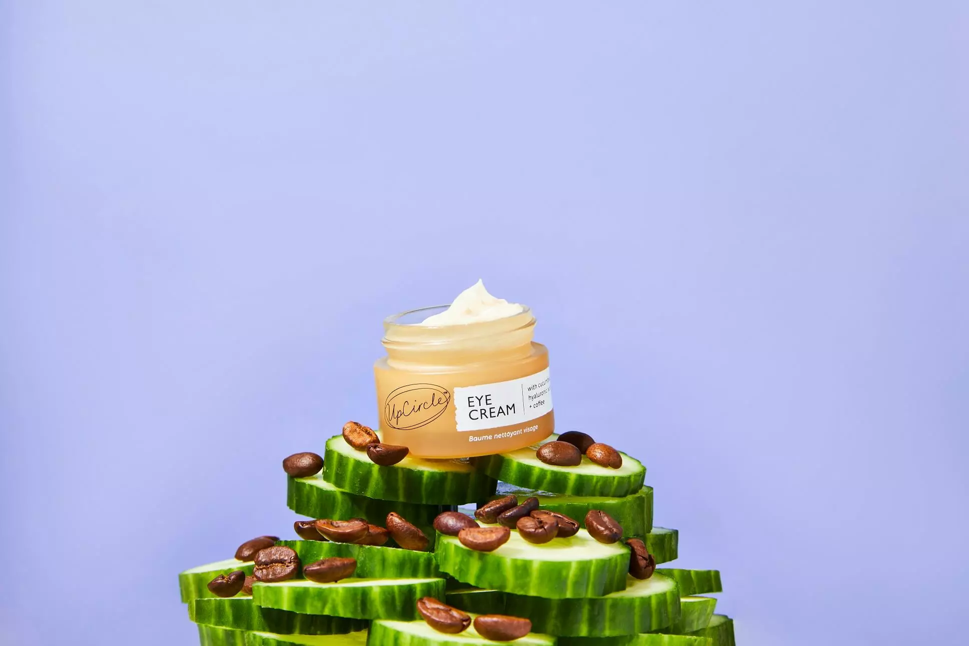 A small jar of UpCircle Eye Cream sits on a stack of sliced cucumbers and coffee beans.