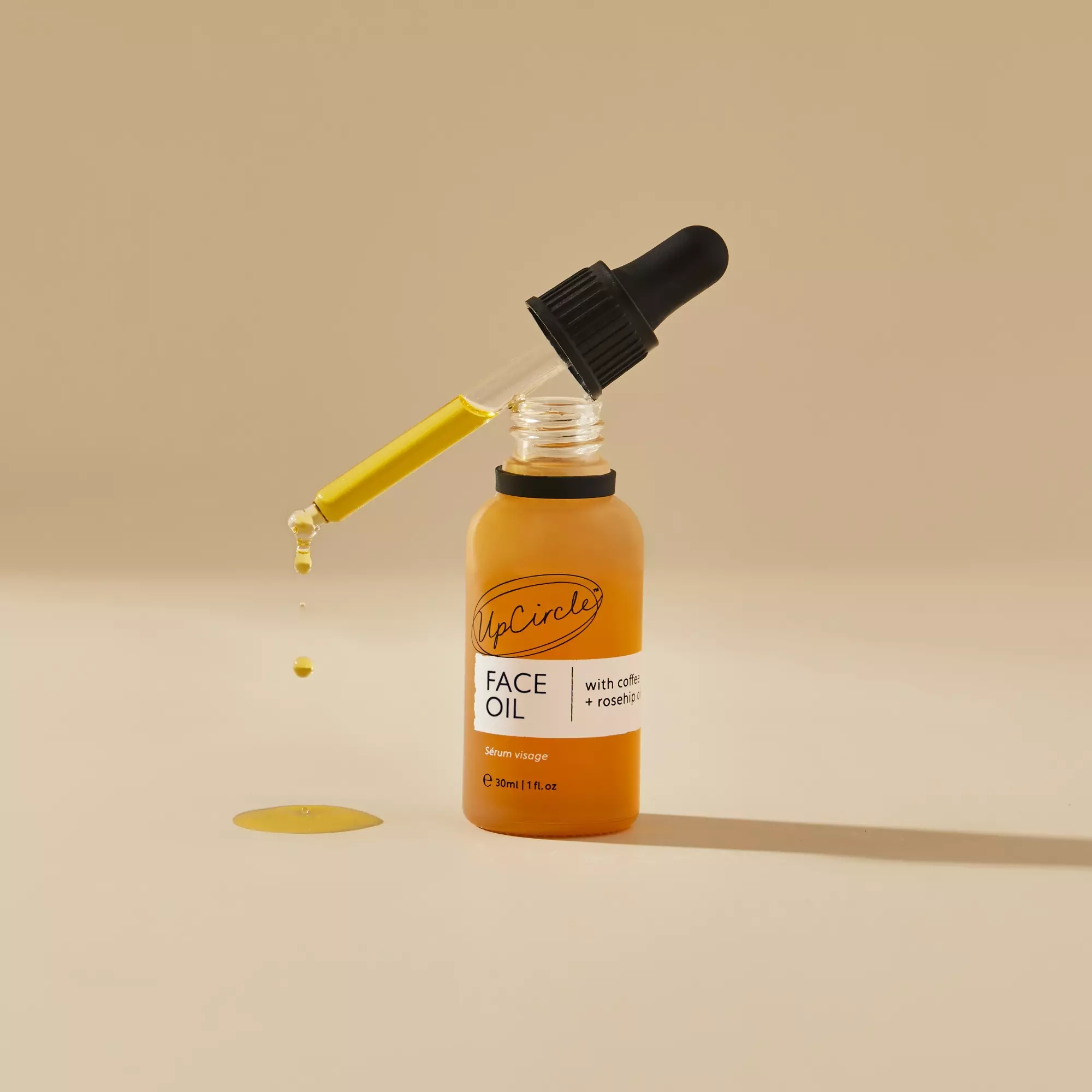 A bottle of UpCircle Face Oil with a dropper dripping yellow oil onto a tan surface.