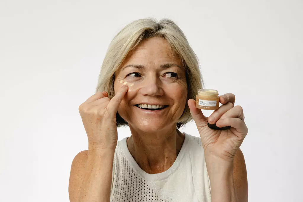 A smiling older woman with short blonde hair holds a small jar while applying eye cream under her eye.