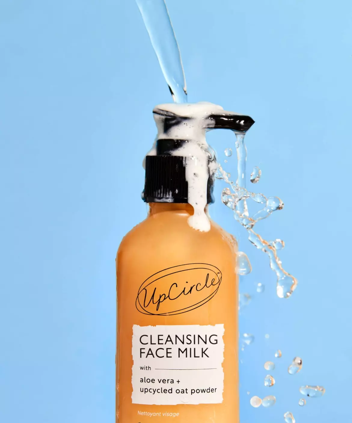 A bottle of UpCircle Cleansing Face Milk with water splashing over its pump against a blue background.