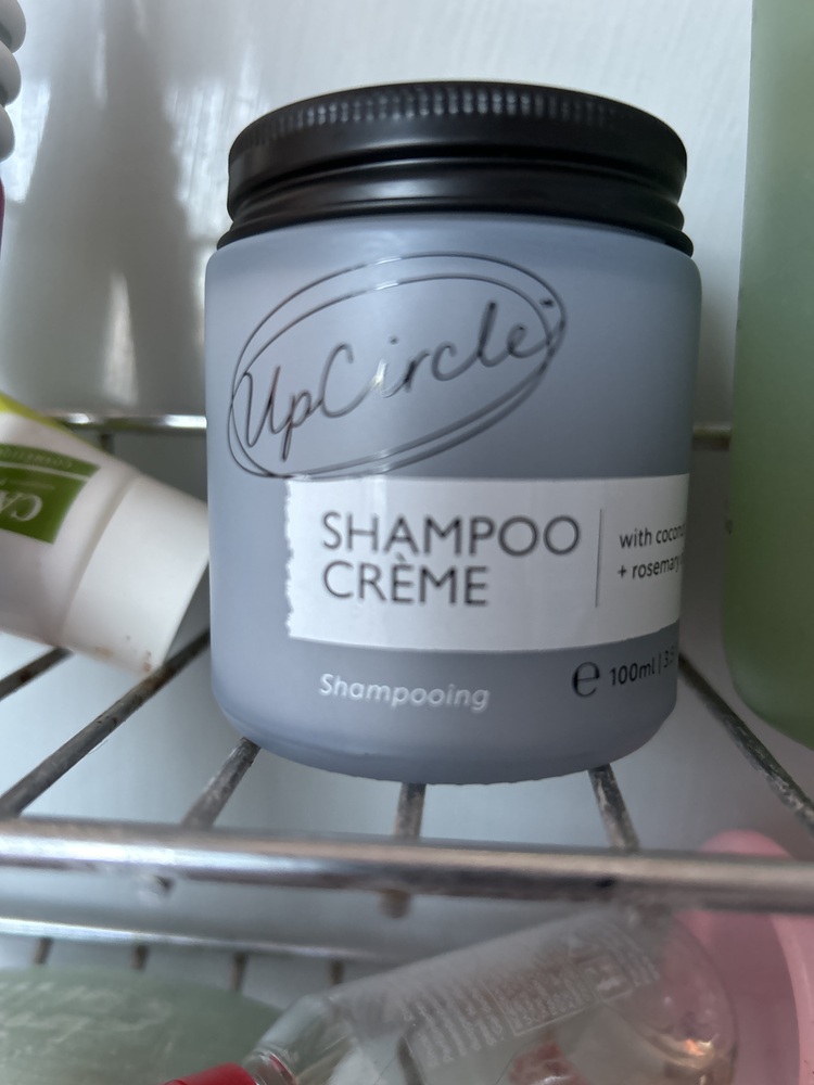 A grey jar of UpCircle Shampoo Crème with a black lid sits on a wire shower rack.