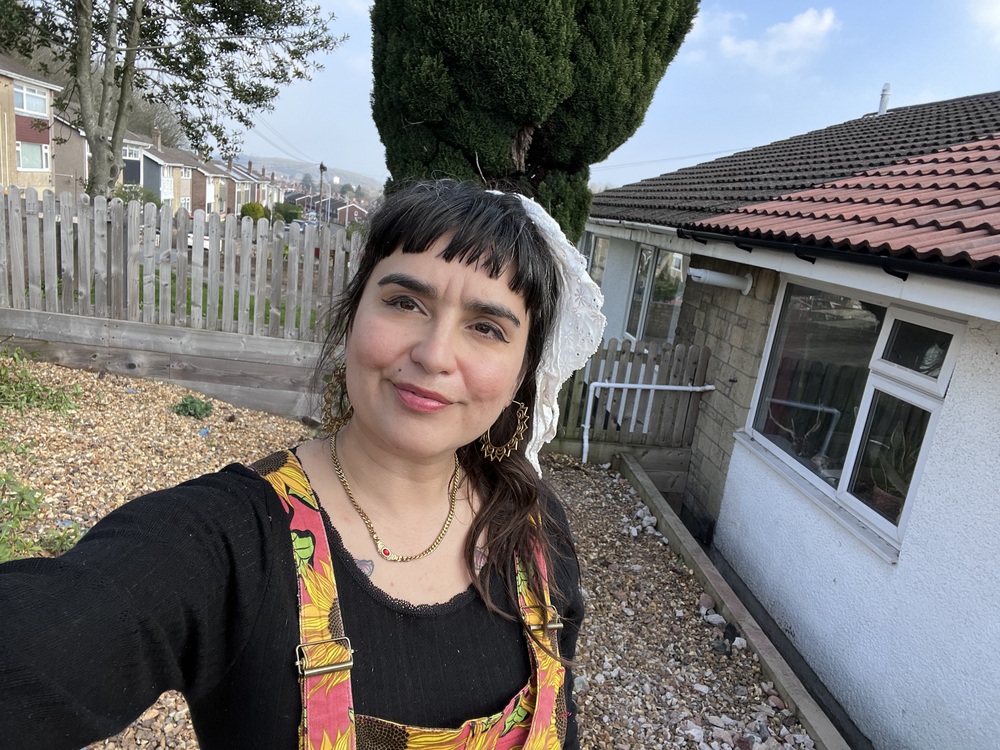 A woman with dark hair and a white lace hair covering takes a selfie in a residential yard.