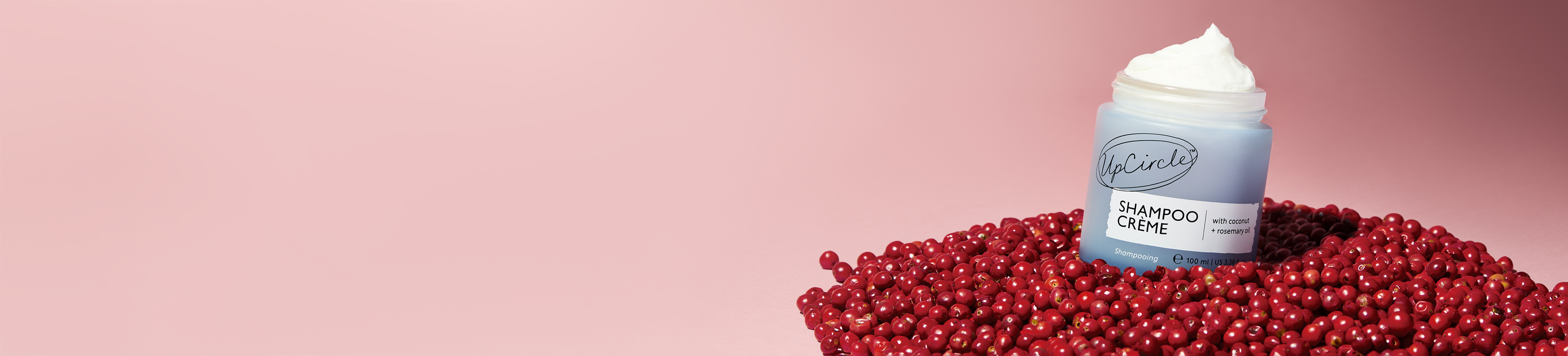 A jar of UpCircle Shampoo Crème sits in a pile of small red berries against a pink background.