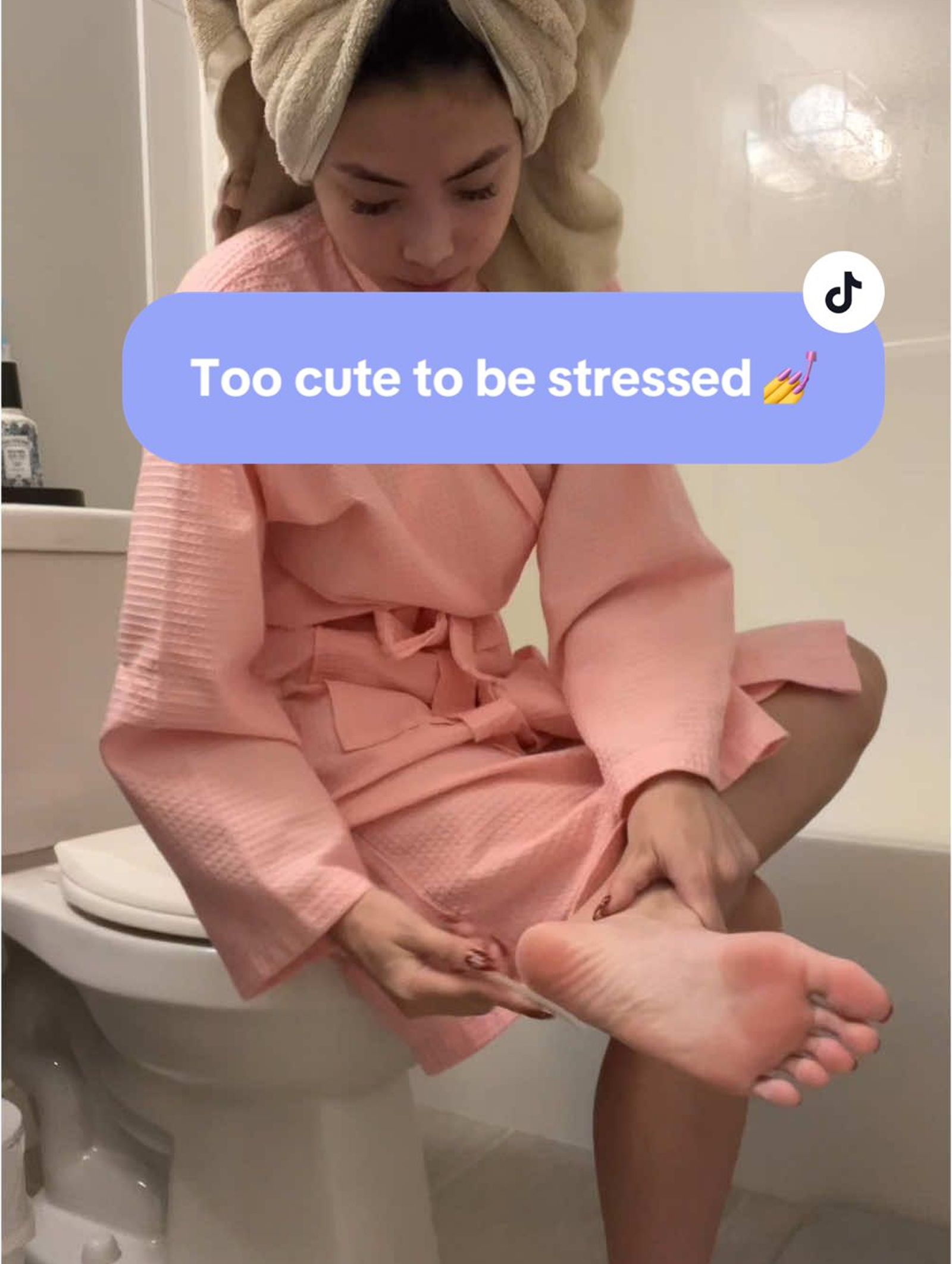Person in a pink robe with a towel on head, sitting on a toilet.