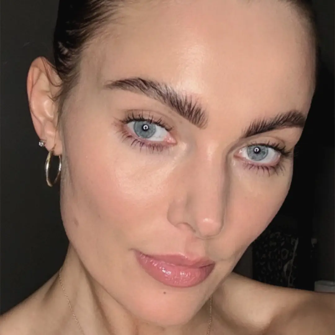 A close-up of a person with blue eyes, prominent eyebrows, and hoop earrings.
