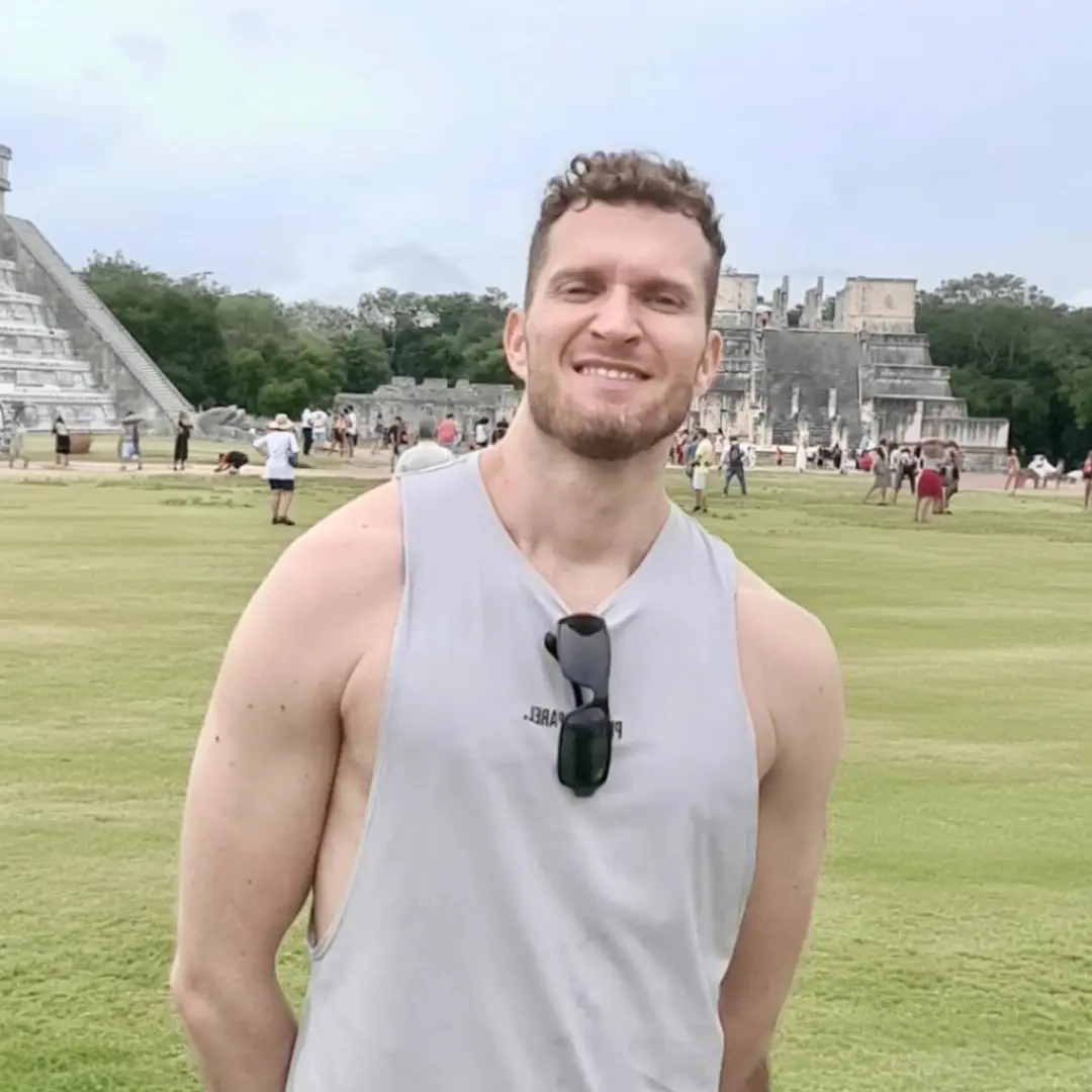 Person smiling in front of an ancient stone pyramid and grassy landscape.