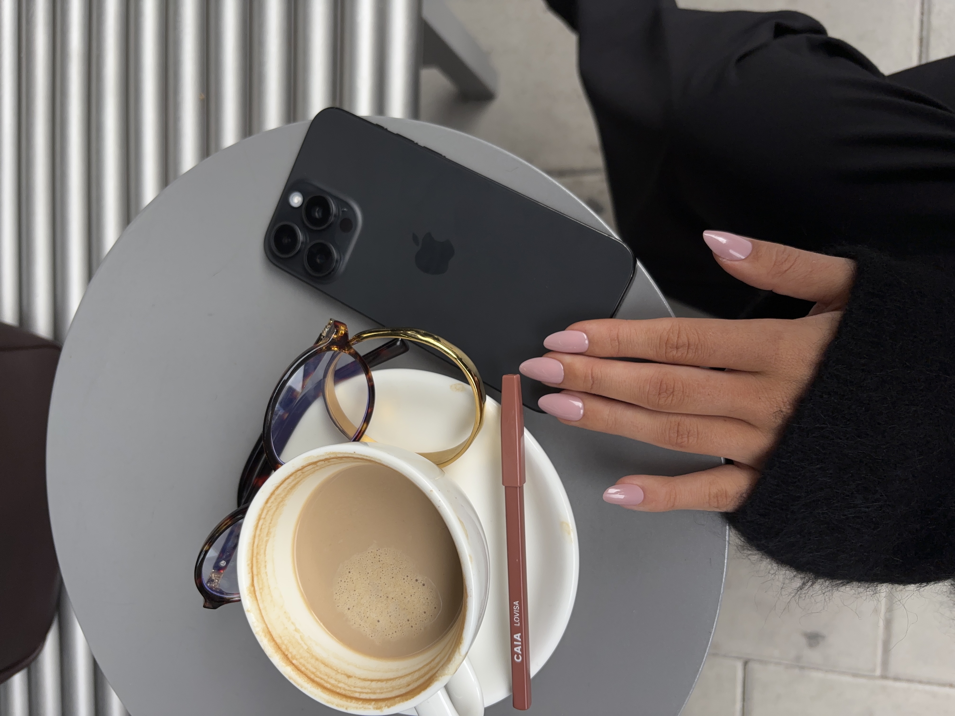 A hand with pink nails rests on a gray cafe table with a coffee, phone, and glasses.