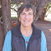 A smiling woman with short brown hair, wearing a blue shirt and a dark vest, stands outdoors.