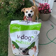 A smiling tan dog stands on a porch behind a large bag of v-dog kind kibble.
