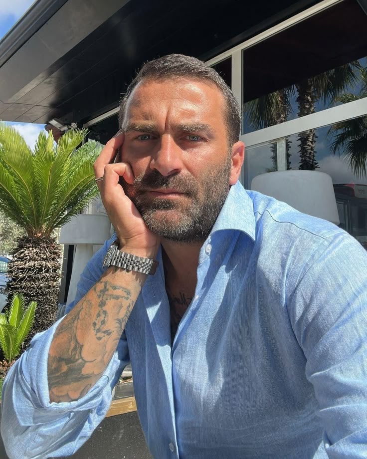 A man with tattoos wearing a blue shirt, sitting outdoors with palm trees in the background.