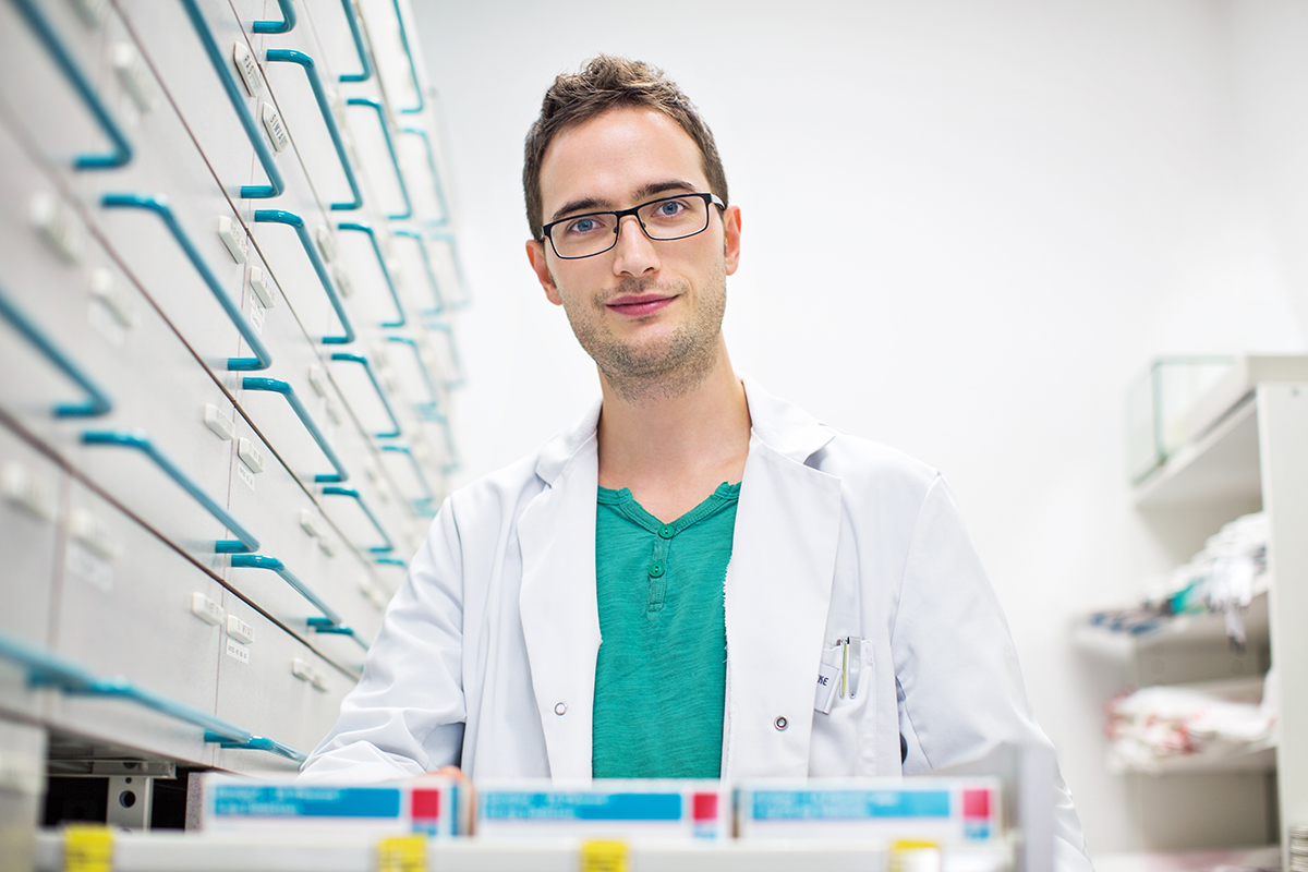 A person in a white coat standing by shelves of drawers in a bright room.