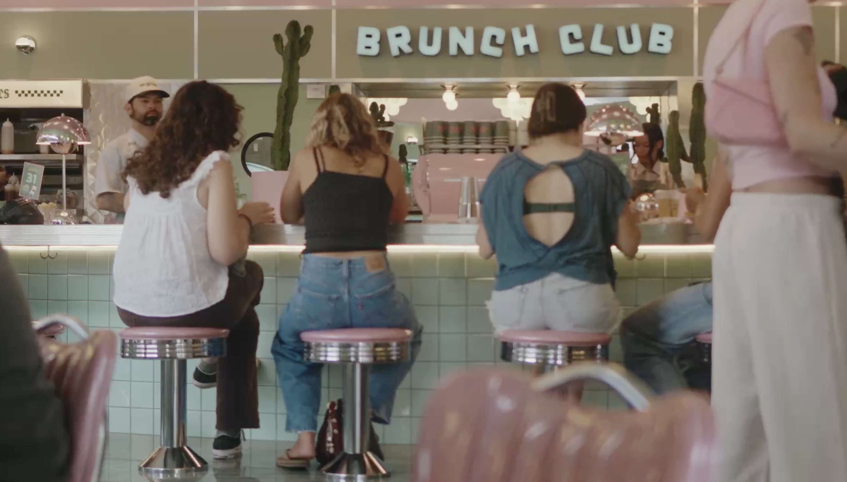 The backs of several women sitting on pink stools at the counter of a retro diner called Brunch Club.