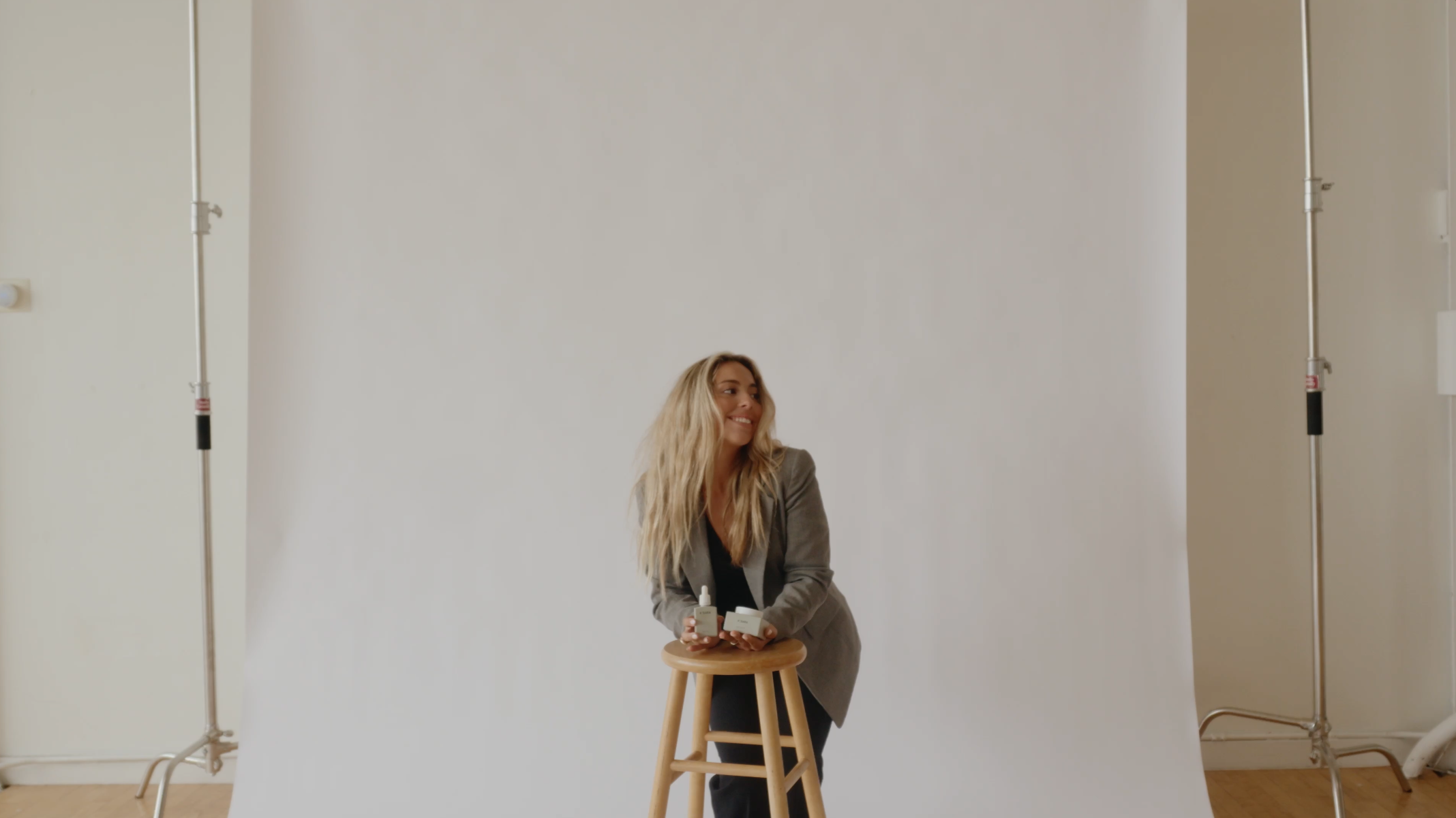 A smiling blonde woman in a grey blazer leans on a stool in a photo studio, holding products.
