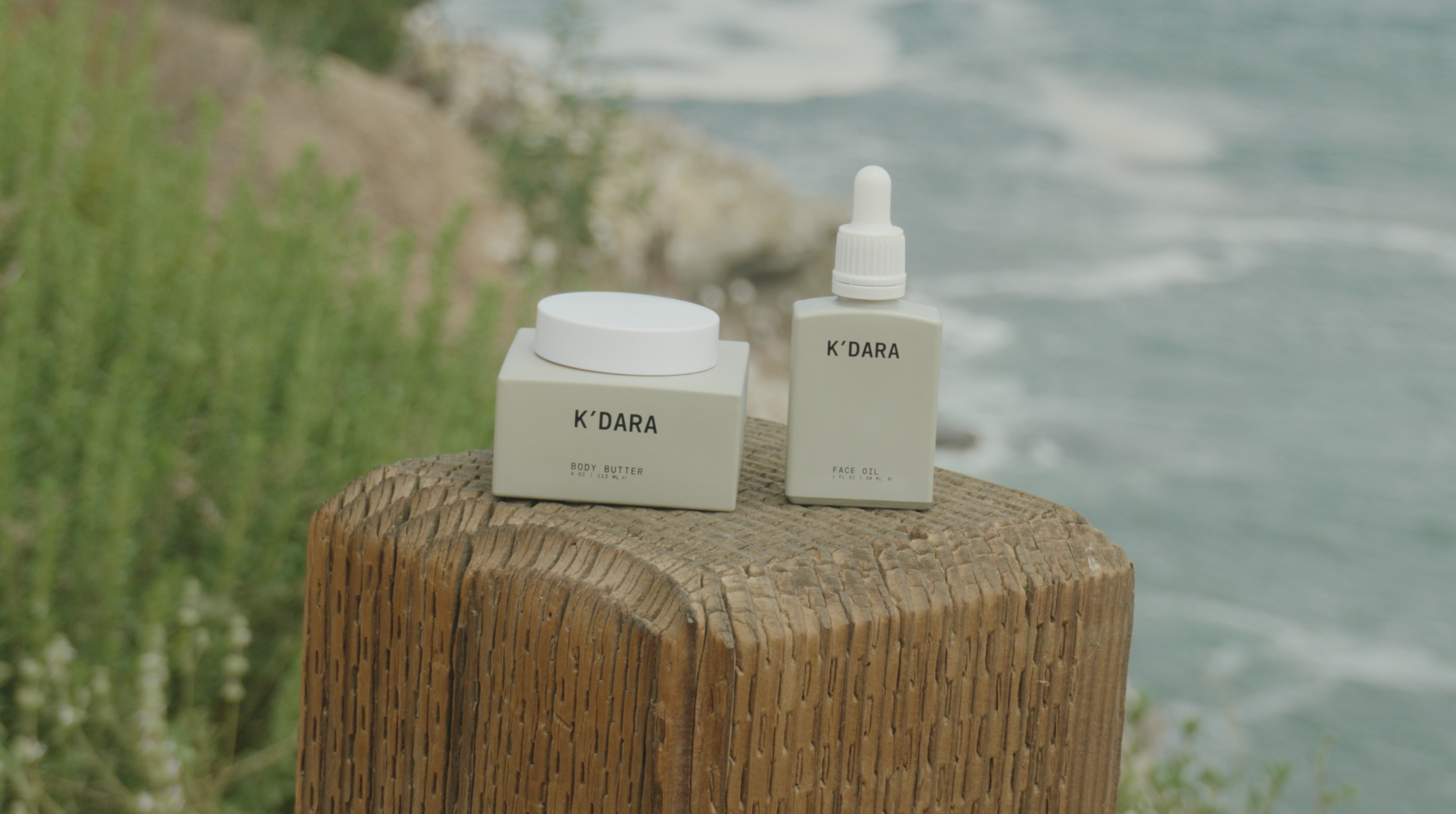 K'DARA Body Butter and Face Oil sitting on a wooden post with the ocean in the background.