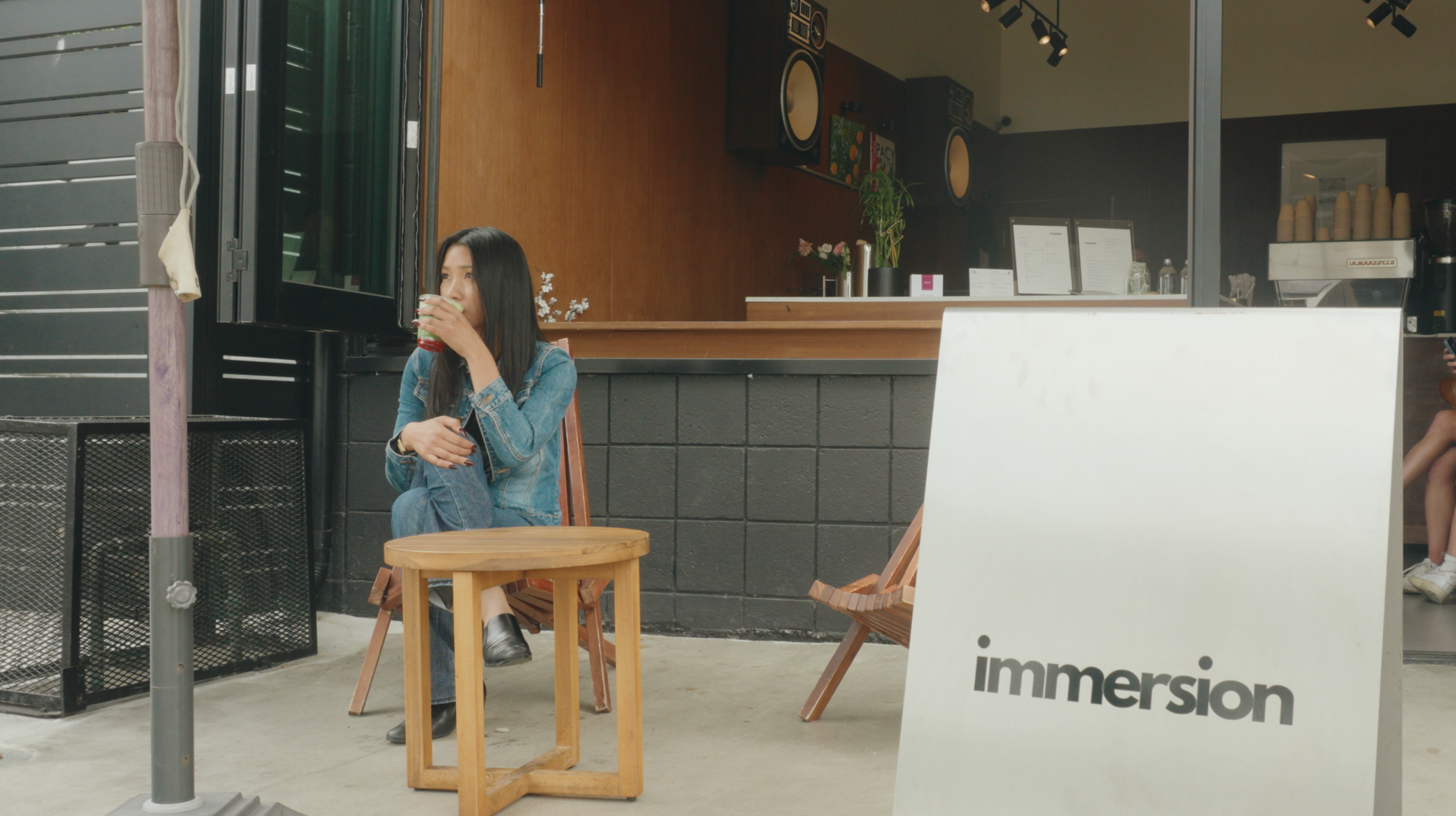 A woman in a denim jacket sips a drink while sitting at a small wooden table outside a cafe.