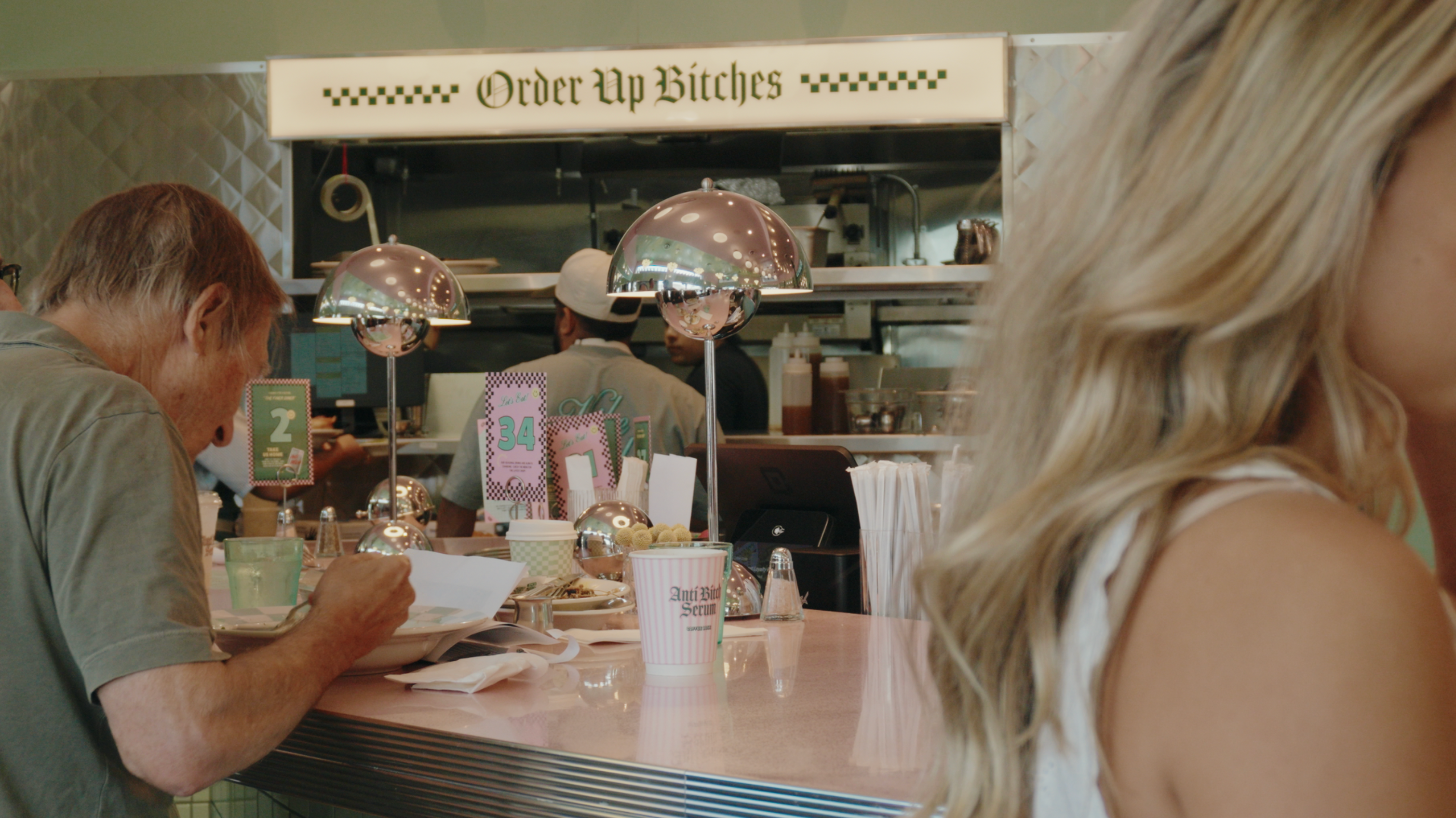 People sit at the counter of a retro-style diner with a sign that reads 'Order Up Bitches'.