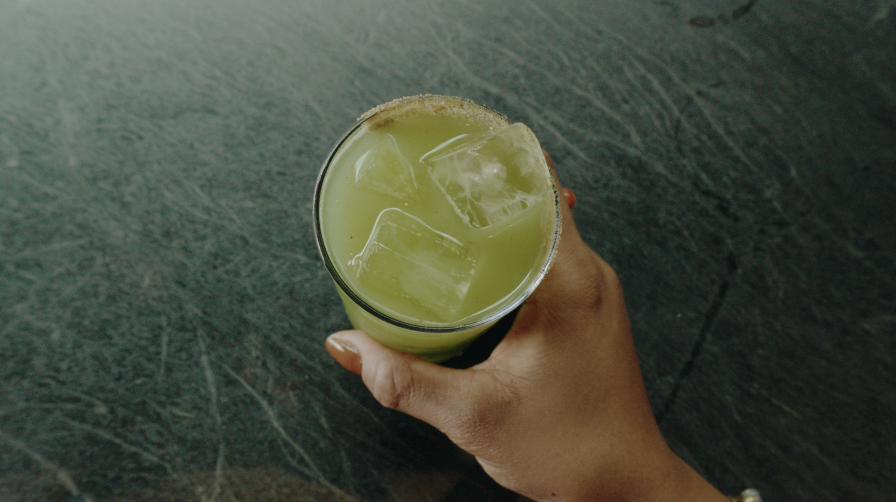An overhead shot of a hand holding a green cocktail with ice and a garnished rim.