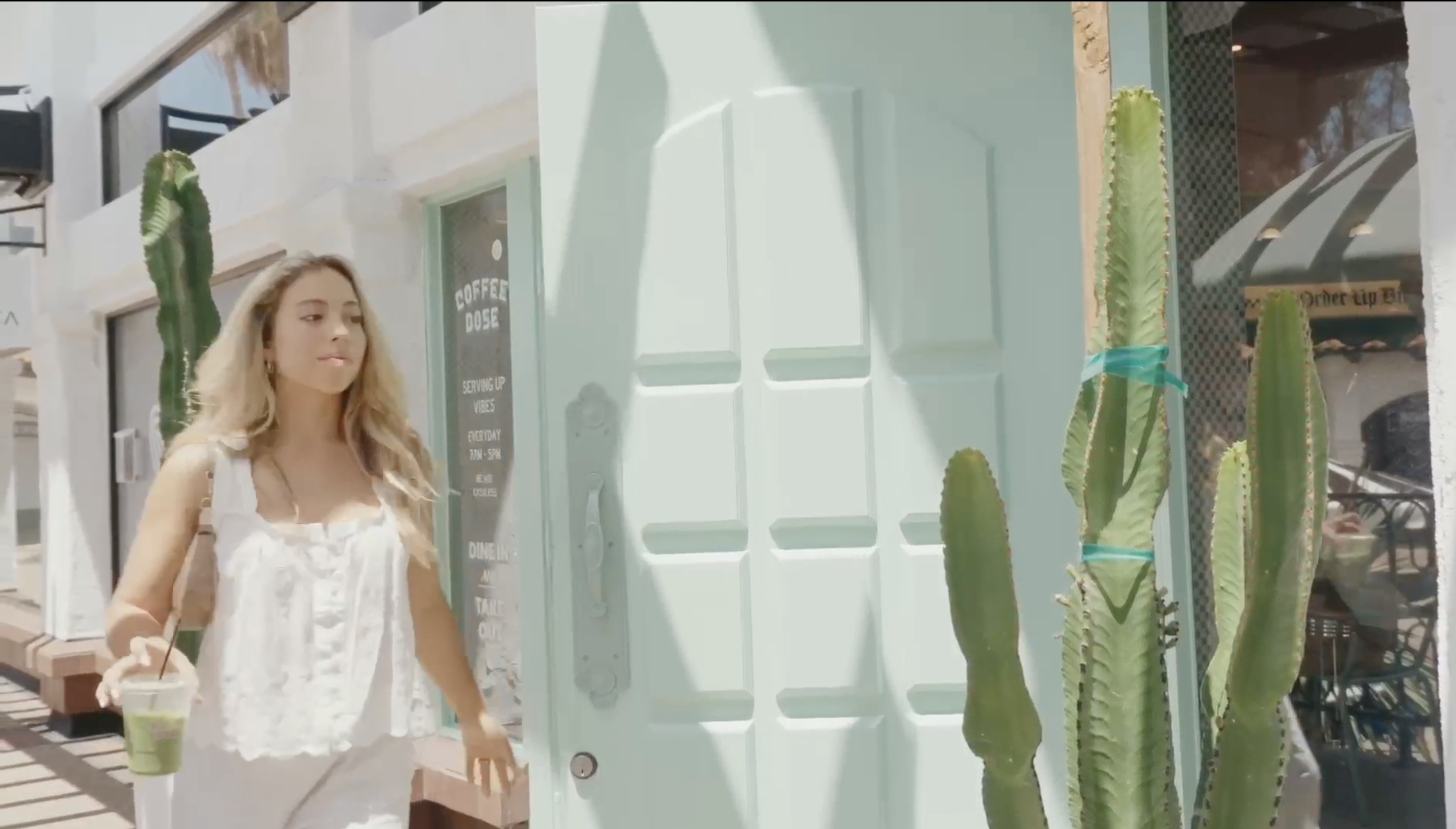 A woman with a green drink walks out of a coffee shop next to a light blue door and cacti.