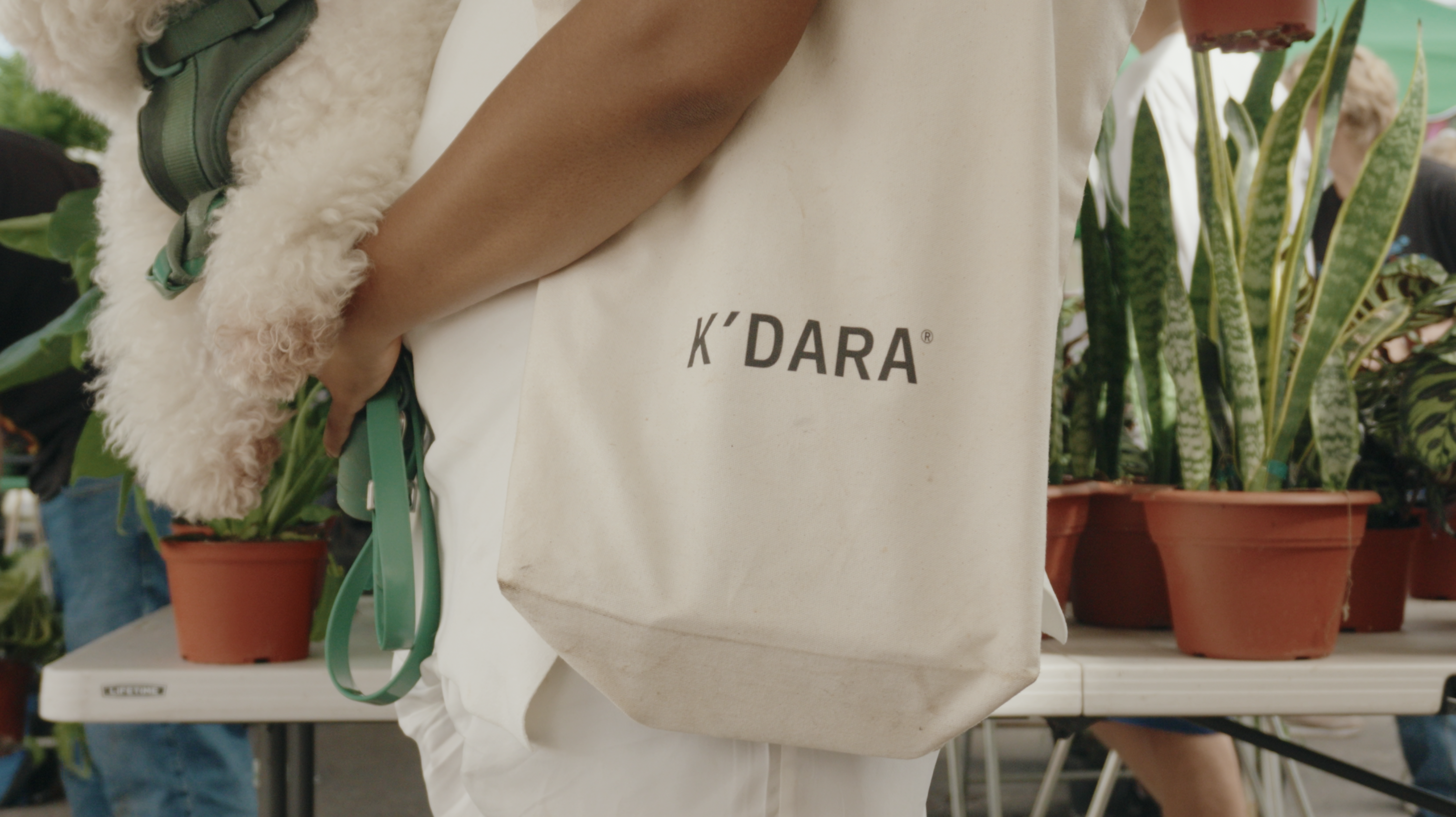 A person holding a white fluffy dog and a K'DARA tote bag next to a table of plants.