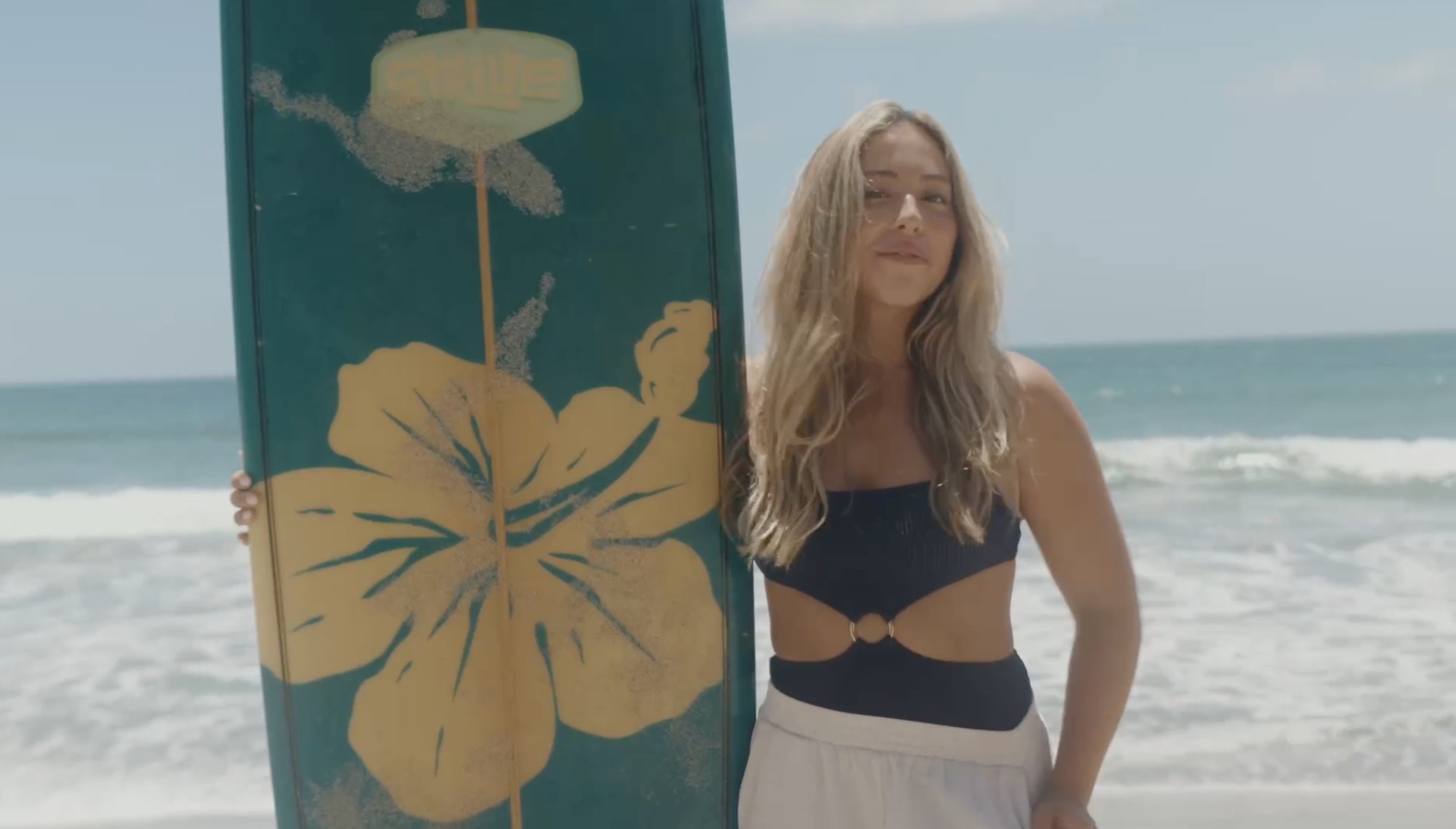 A woman stands on a beach holding a surfboard with a large yellow flower design.