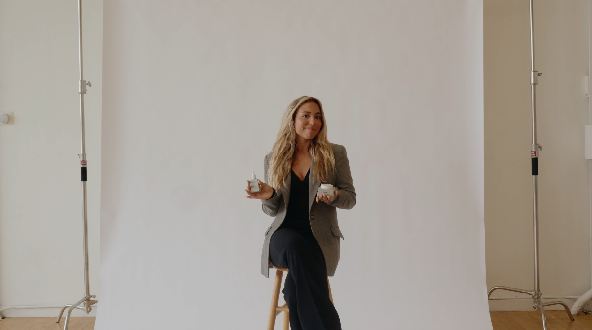 A woman in a blazer sits on a stool, holding two skincare products in a photo studio.