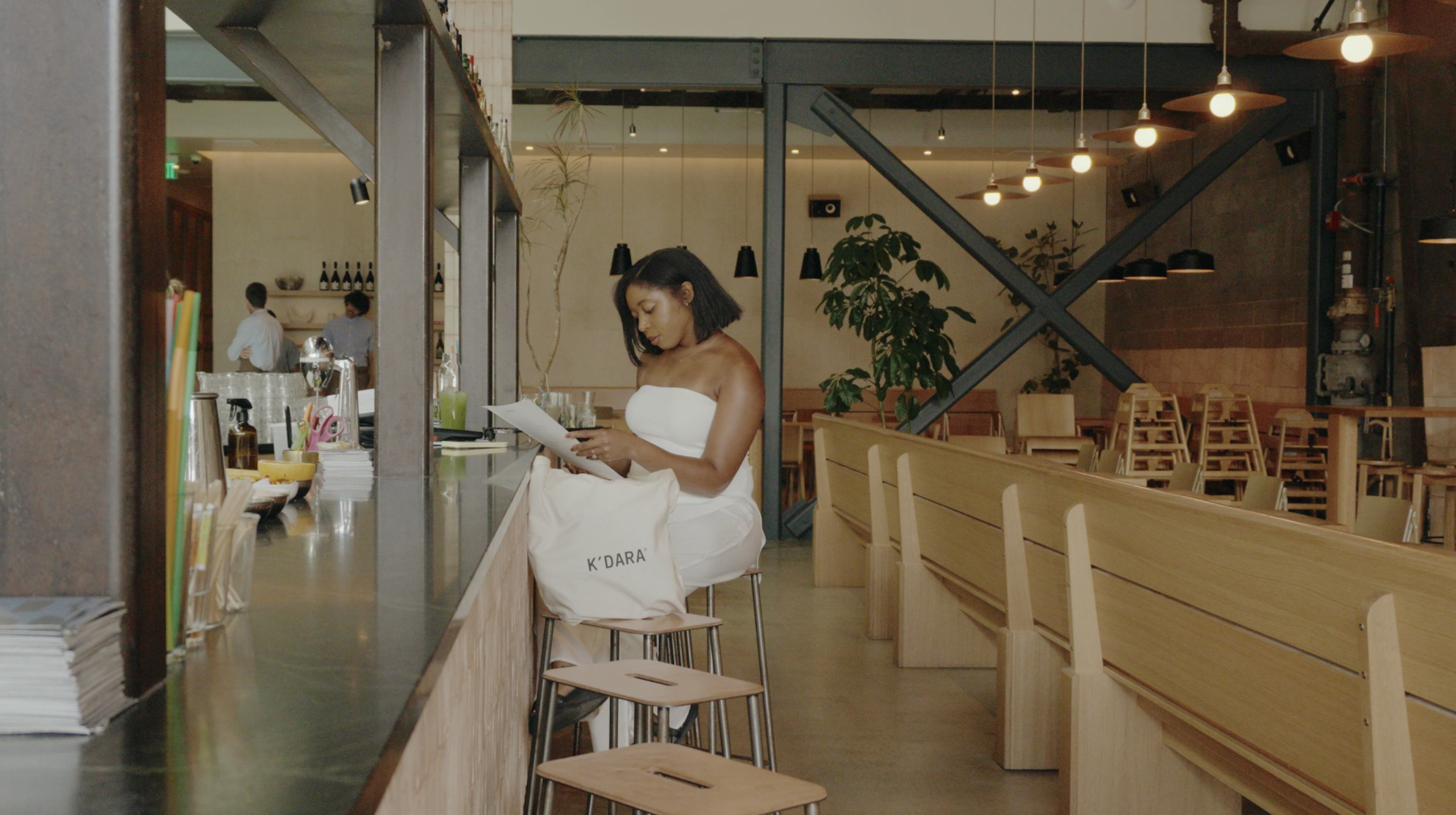 A woman in a white outfit sits at a modern restaurant counter reading from a piece of paper.